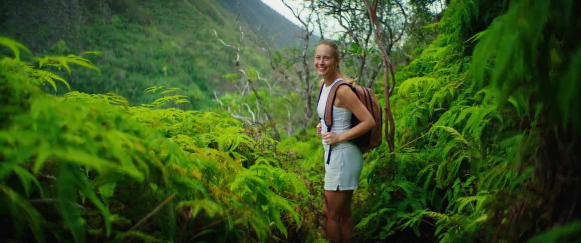 A woman with a backpack hikes through a vibrant green forest with a mountain landscape in the background