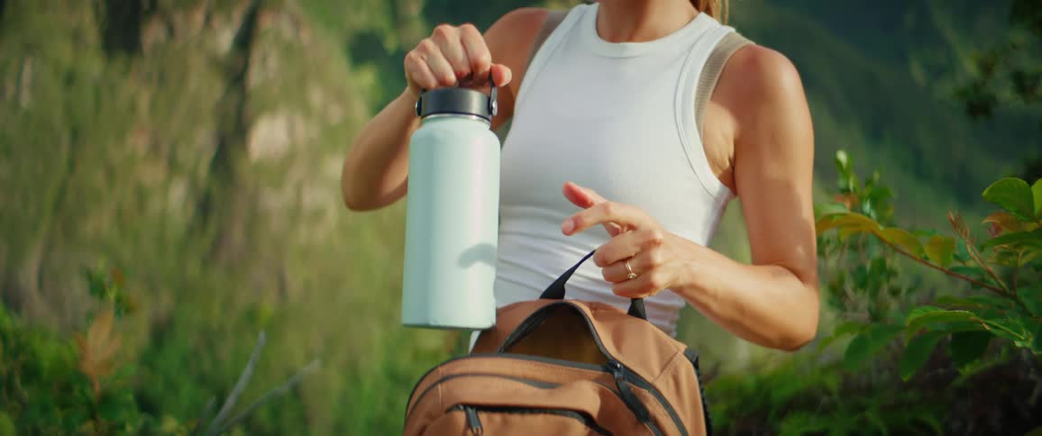 A woman with a ponytail and a white tank top stands on a trail with a green mountain landscape behind her taking a light blue water bottle from a backpack unscrewing the cap and taking a drink