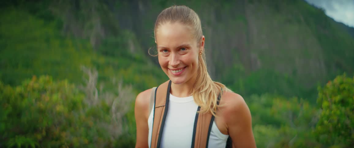 A blonde woman wearing a white tank top and a backpack is standing outdoors she is smiling and looking directly at the camera she appears to be hiking with lush green hills visible in the background