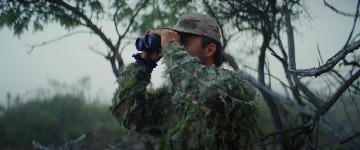 Camouflaged hunter scans the misty woods with binoculars