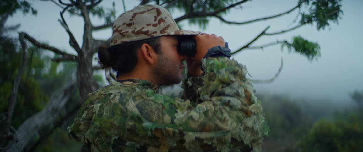 A man dress entirely in camouflage looks through a pair of binoculars
