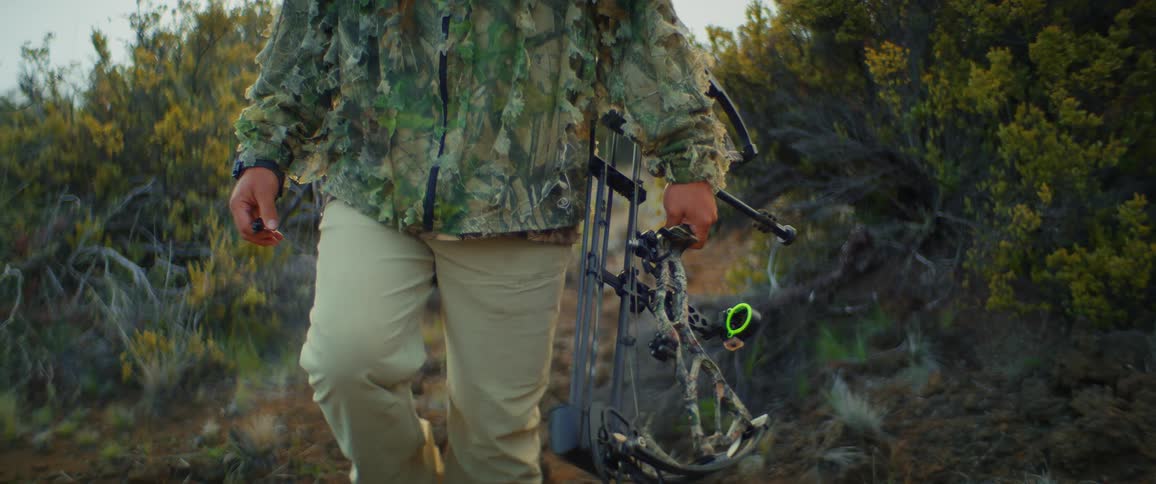 A man walking on a bath through the woods wearing camouflage clothes and carrying a professional shooting bow