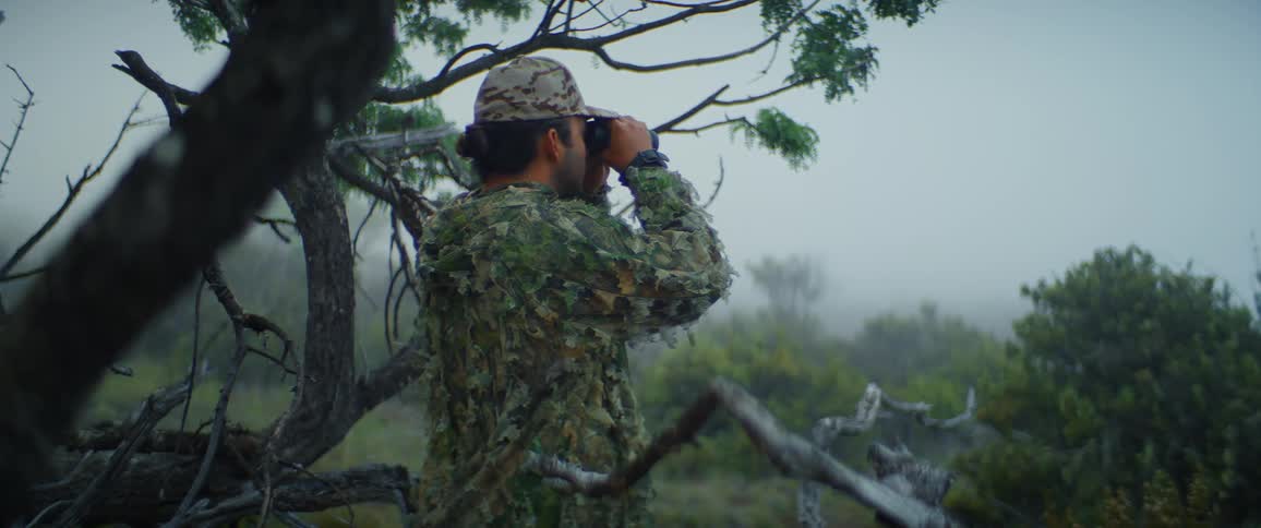 Man in camouflage clothing and cap looks through binoculars in a wooded foggy area