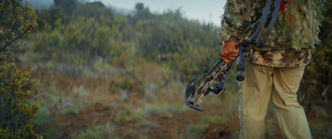 A man wearing camouflage walks uphill while carrying a hunting bow