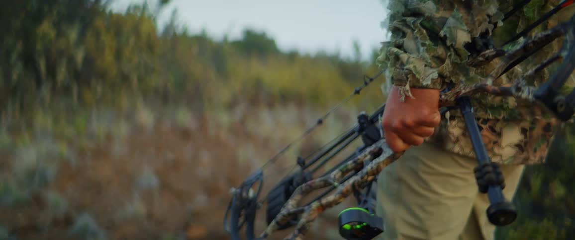 Tight tactile shot of a person carrying a camouflaged compound bow while moving through scrubby terrain