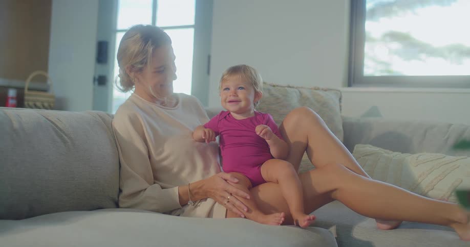A blonde mother and her baby sit on a gray sofa with the baby smiling while sitting on the mothers lap before the mother gently pats the baby on the head