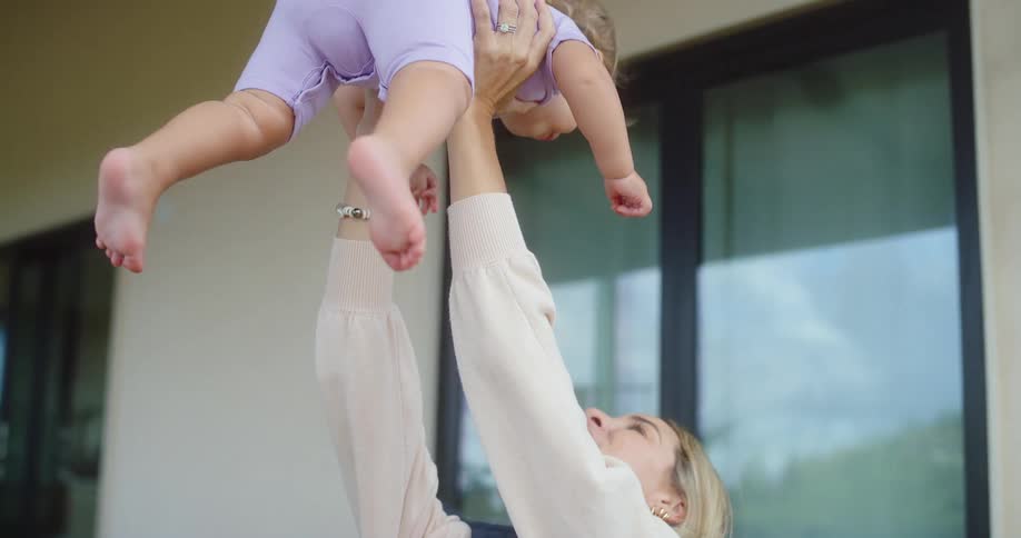 A smiling woman with blonde hair holds a baby in her arms and lifts it up into the air