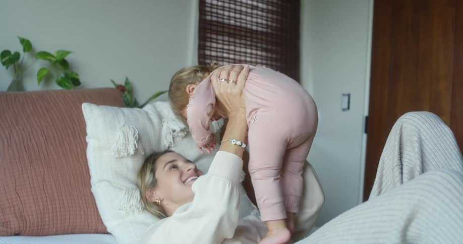 A smiling mother with blonde hair lies on her back in a bed with white pillows and sheets holding a young child in the air with her arms and legs