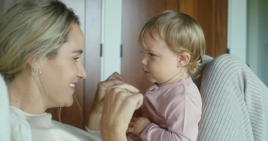 A mother holding her infant on her lap while playing with him as she lies on the bed at midday