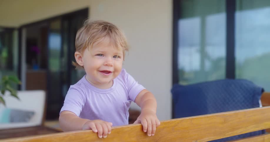 A smiling toddler leans on a wooden railing outside a modern home