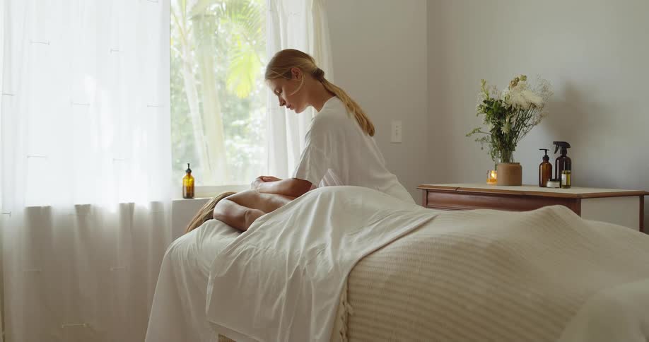 A masseuse with blonde hair and a white top rubs the back of a client who is lying face down on a massage table covered in a white towel with light streaming through a window behind them