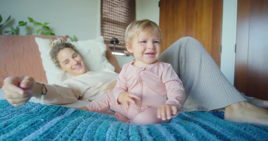 Mother lying on the bed smiles while her child crawls to grab a stuffed toy