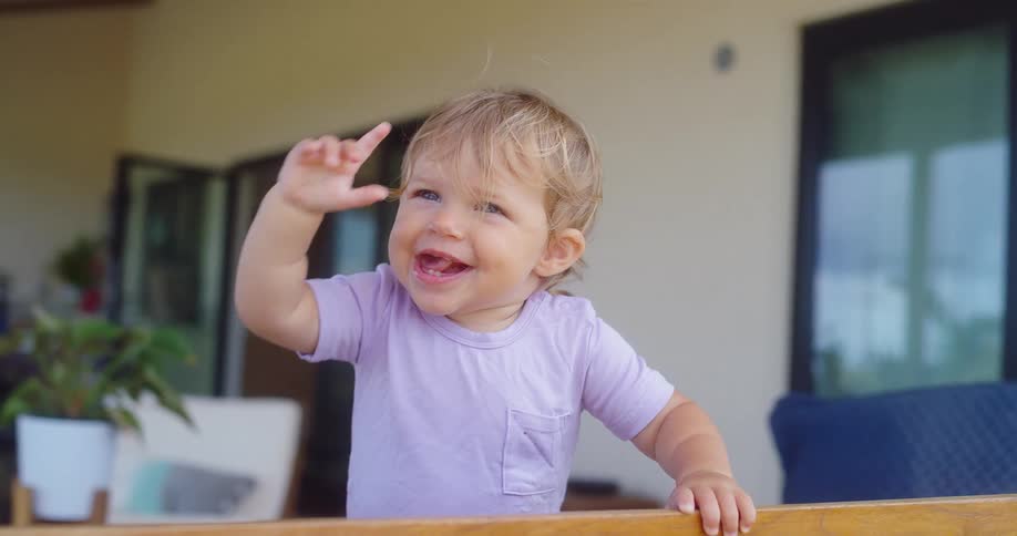 A smiling toddler waves while standing near a railing indoors