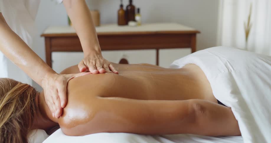 A massage therapist with a white top uses both hands to rub oil on the back of a client with blonde hair who is lying face down on a massage table covered in white sheets