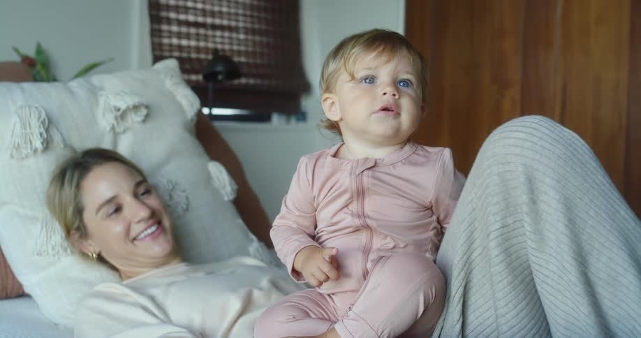 A blonde woman lies on a bed smiling at a child with blue eyes who is sitting next to her the child looks offcamera as the woman laughs in the background