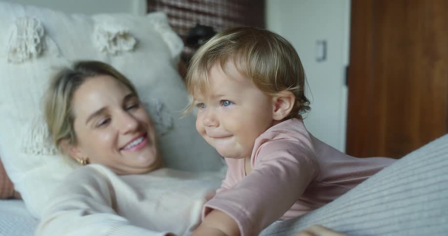 A mother and her baby are playing together on a bed the baby has blonde hair and blue eyes and is looking up at the mother who is smiling warmly at the baby the mother is lying down with her head on a pillow