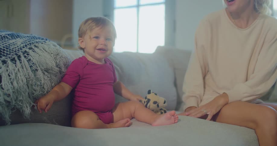 A happy baby in a red onesie with a toy leopard on their lap sits on a gray sofa next to a smiling woman and looks directly at the camera