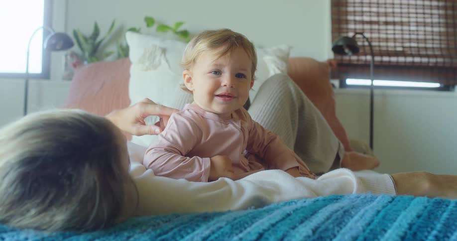 A baby with blonde hair and a pink onesie sits on a persons stomach looking up and smiling before turning to look at the camera