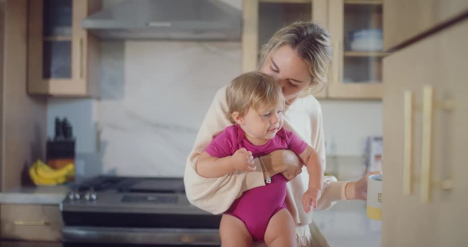 A blonde woman holds a young child in her arms while standing in a kitchen the child wearing a pink onesie looks around while the woman smiles down at them