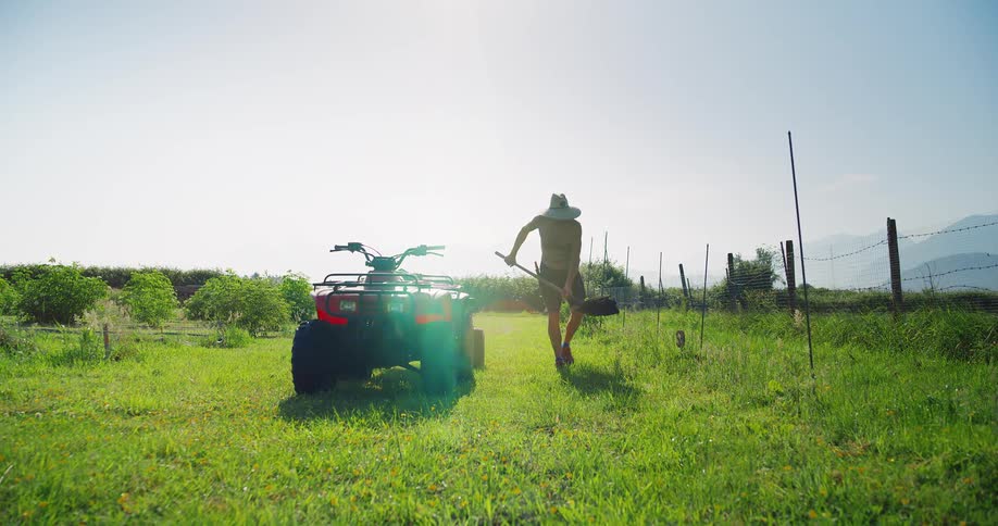 A farmer wearing a sunhat uses a shovel to scoop compost from an atv and spread it on the soil in a grassland at midday