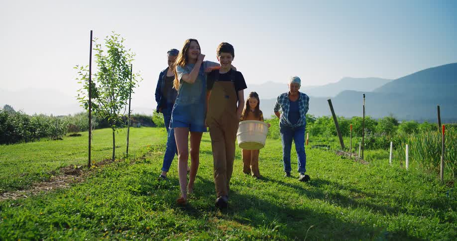 In a sunlit field a group of four people walk toward the camera a young man wearing brown overalls walks arminarm with a young woman in denim shorts and a jacket behind them an older man and a young girl carry buckets with mountains visible in the