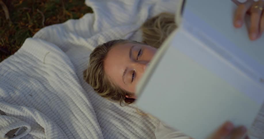 Person lying on a white blanket reading a book outdoors