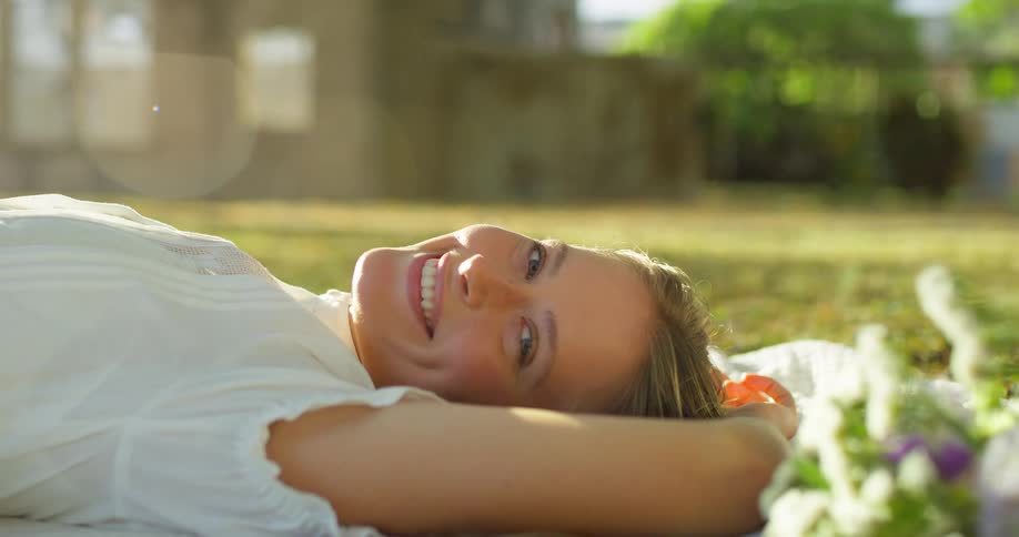 Smiling woman laying with arms over her head on white blanket on grassy lawn