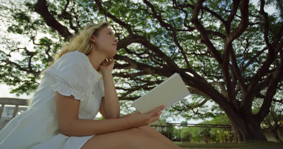 The woman holds a book while sitting under the tree