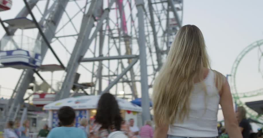 A blonde woman in a white tank top and blue jeans is walking through a fairground she turns to face the camera smiles and then turns back around to continue walking towards a ferris wheel