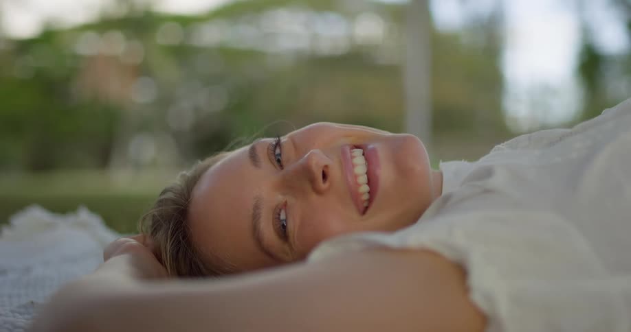 A blonde woman is shown lying on her side outdoors looking into the camera she smiles then closes her eyes appearing relaxed