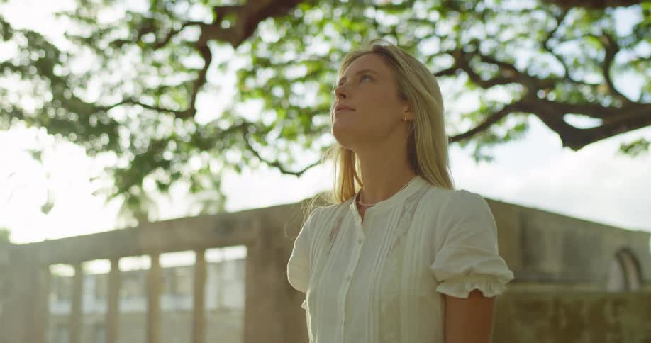 A blonde woman stands outside looking up at the sky she then turns her head to the side looking offcamera she is wearing a white dress with a large tree and a building visible in the background
