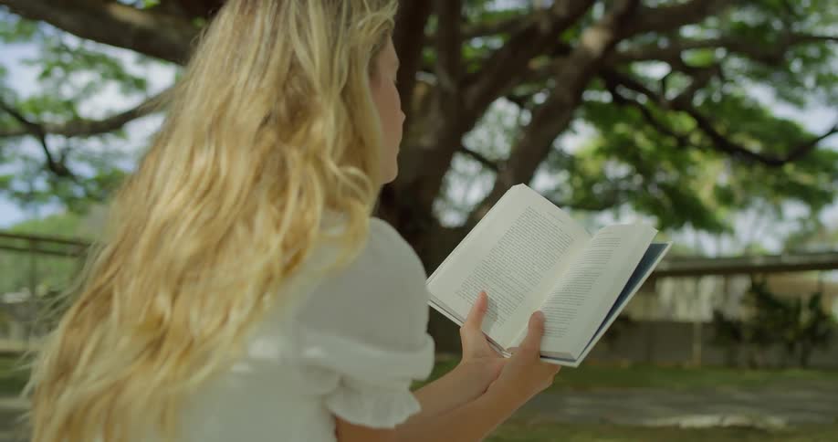 Blonde girl sits outdoors and reads while a large tree stands in the same field