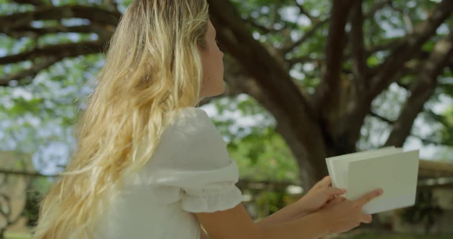 Blonde woman sitting outdoors under the wide canopy of a large tree reads from a book