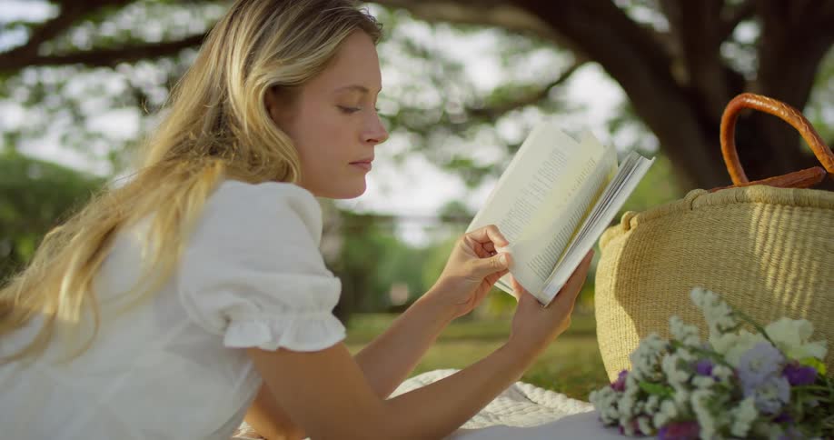 A blonde woman in a white dress is seen relaxing under a large tree she is lying on a blanket and reading a book