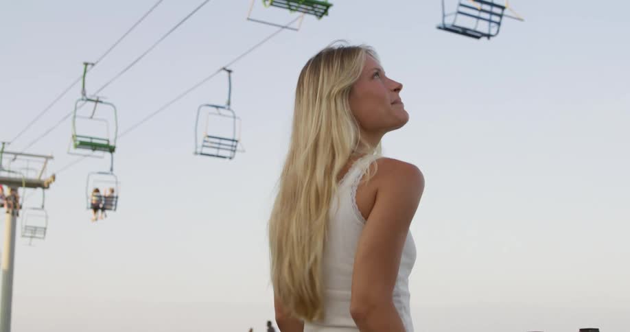 A blonde woman in an amusement park watching people ride a ski lift during summer at midday smiling