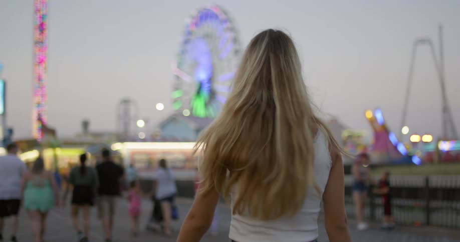 A blonde woman smiles while walking around a carnival at dusk