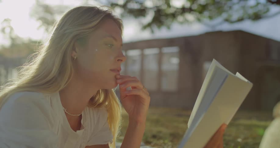 A blonde woman sits outdoors in the sun wearing a white shirt and a necklace as she holds an open book and reads with a concentrated expression on her face