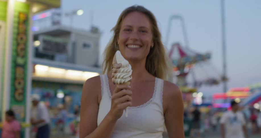 A woman happily eats an ice cream cone at a carnival
