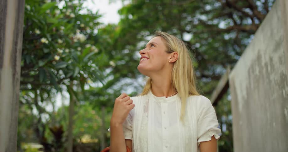 A woman with blonde hair and a white button up top with ruffles on the sleeves walks outdoors holding a brown bag looking up with a smiling expression