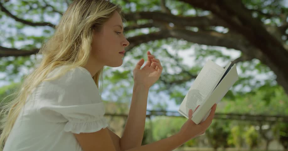 Blonde woman wearing a white dress sitting down outdoors on a park next to trees reading a book during day time