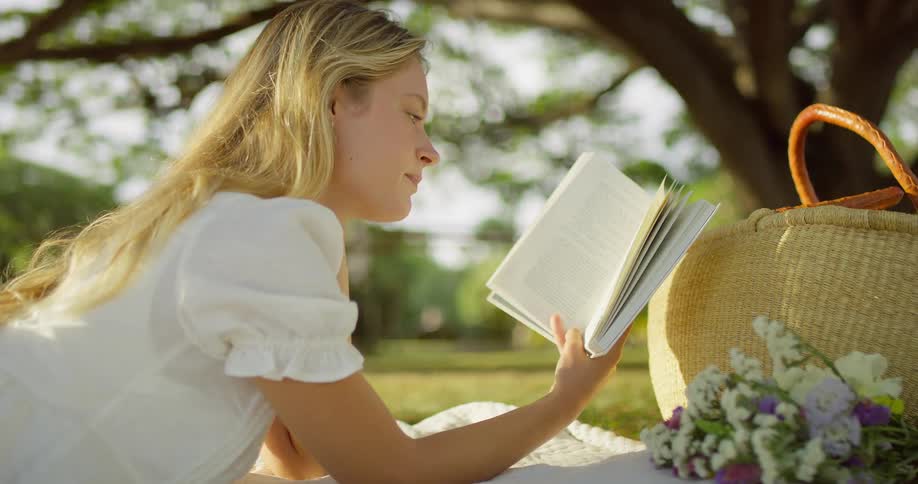 Woman lying outdoors reads a book beside a basket and flowers
