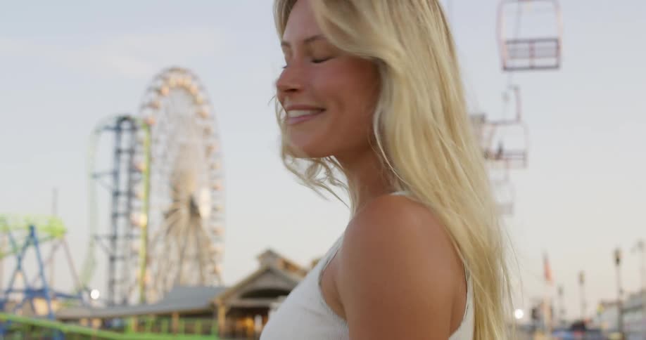 A happy woman with long blonde hair is seen smiling at the camera and then turning to walk away in an amusement park with various rides visible in the background