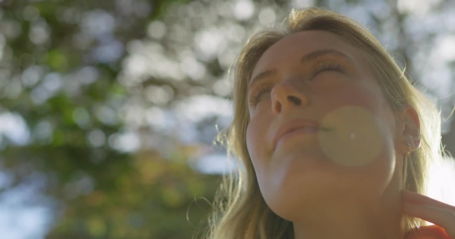 A blonde woman with blue eyes looks up with a thoughtful expression and a hand resting on her neck before smiling and looking to her left
