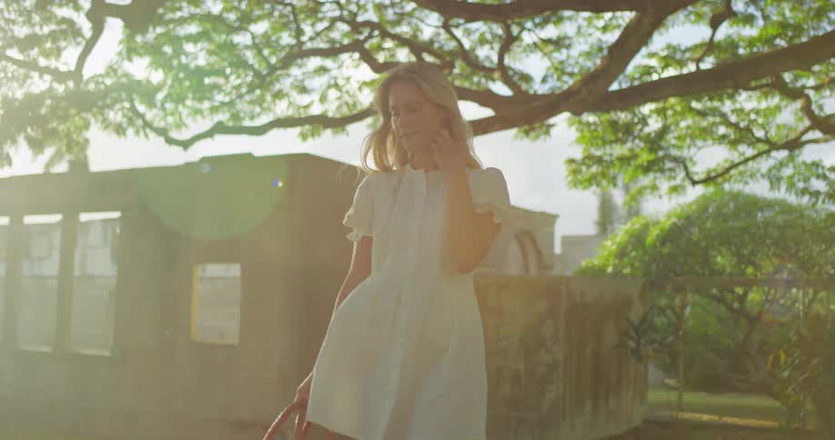 A young woman in a white dress leisurely walking towards the camera holding a basket of flowers as the bright golden sun flares through the trees behind her