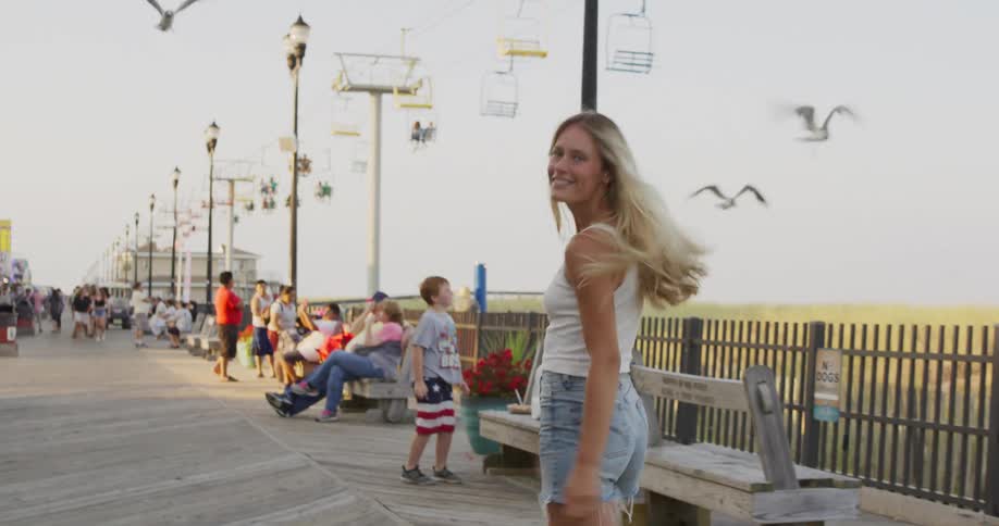 Woman walking on a busy seaside boardwalk with people sitting on benches and a cable car in the background