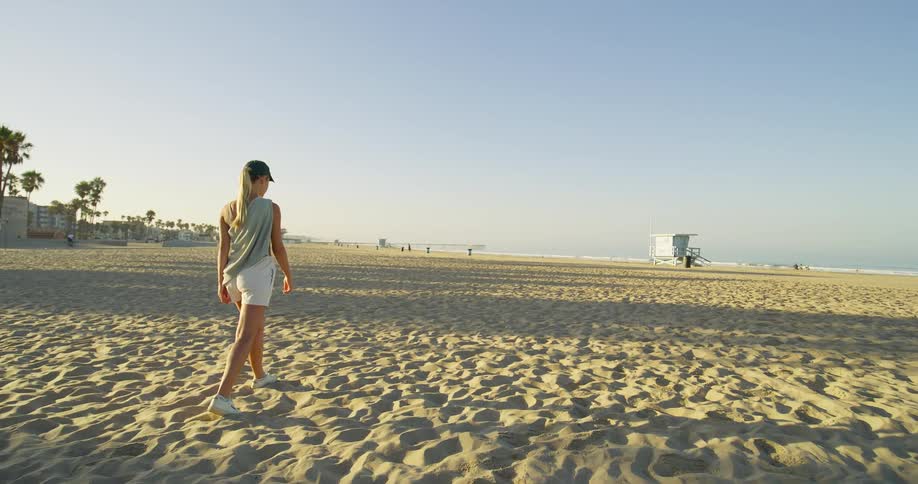 The video shows a wide shot of a young woman walking on a sandy beach she is wearing shorts a loose top a baseball cap and sneakers a lifeguard tower and palm trees are visible in the background