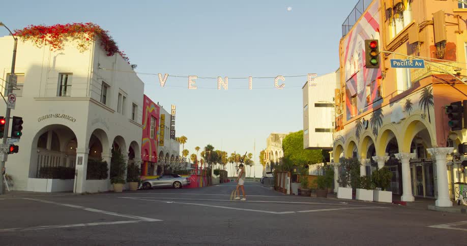 The video shows a wide shot of a street with an archway that reads venice colorful buildings line both sides of the road as a person crosses the street in the distance