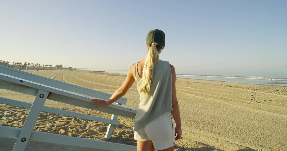 A woman with a long blonde ponytail and a dark baseball cap leans on a white wooden lifeguard stand wearing a white tank top and white shorts as she looks out at the ocean on a sandy beach