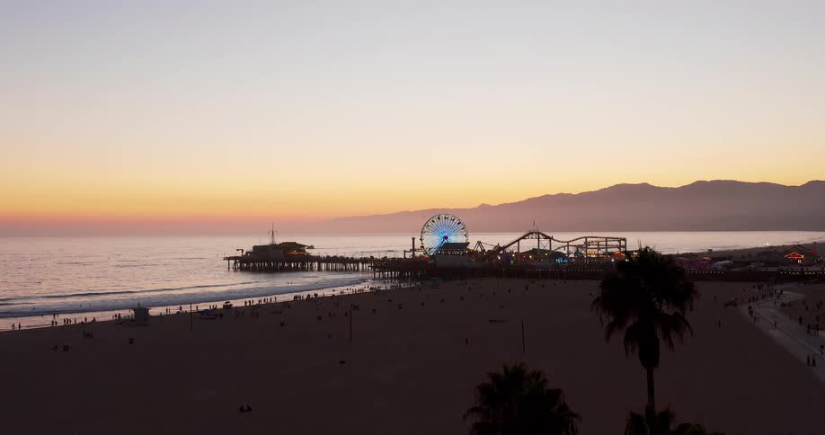An aerial view captures santa monica pier and beach at sunset with the ferris wheel glowing against the colorful sky and mountains in the distance