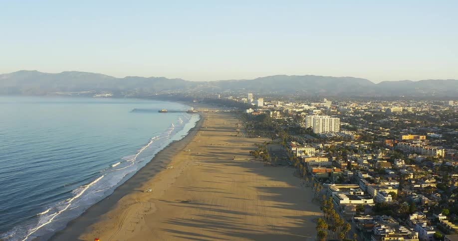An aerial shot shows a wide sandy beach and the ocean on the left with a sprawling cityscape and distant mountains on the right all under a clear blue sky